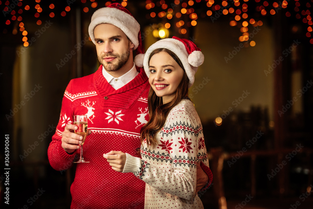 happy young couple in santa hats looking at camera while holding champagne glass and sparkler happy young couple in santa hats looking at camera while holding champagne glass and sparkler