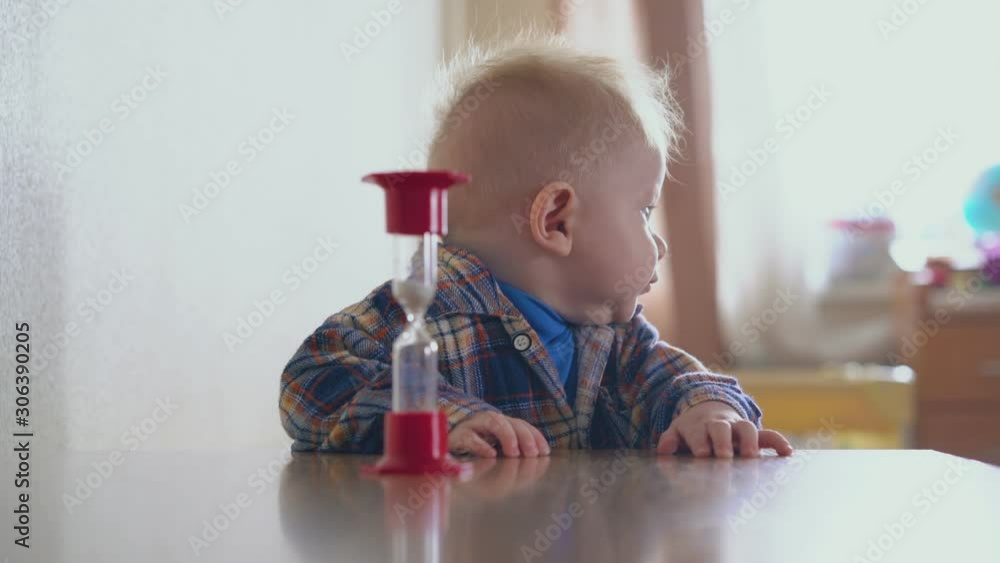 adorable funny baby in checkered shirt stands at table and mommy puts sandglass in light room close view