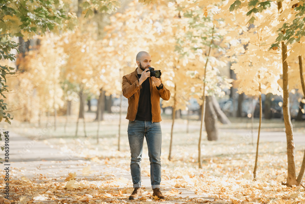 Bald brutal photographer with a beard in a suede leather jacket, blue shirt, jeans, and Chelsea boots goes to take a picture in the park in the afternoon Bald brutal photographer with a beard in a suede leather jacket, blue shirt, jeans, and Chelsea boots goes to take a picture in the park in the afternoon