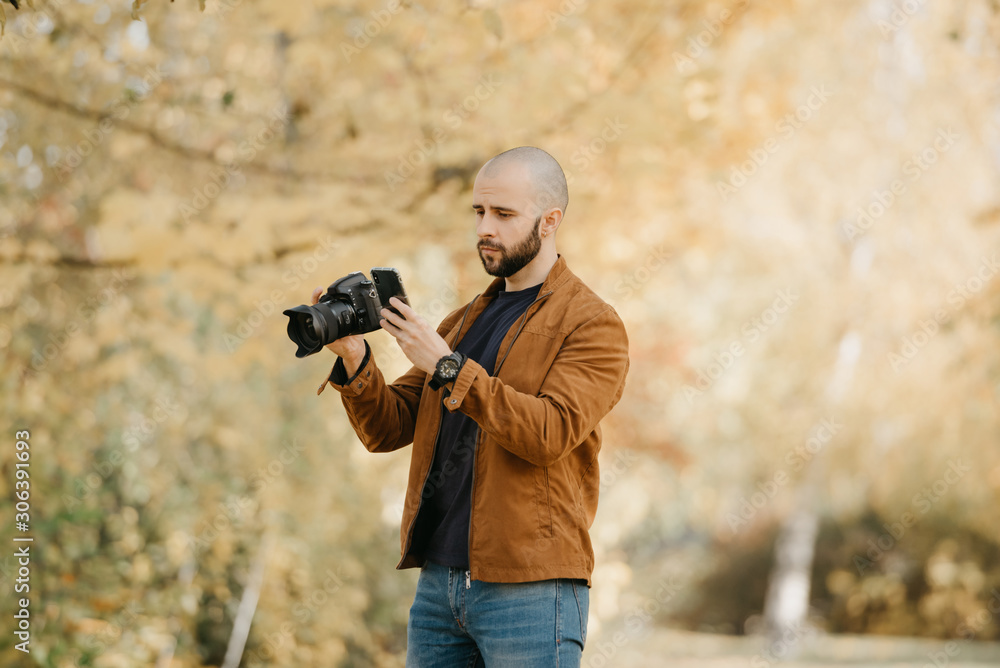 Bald stylish photographer with a beard in a suede leather jacket, blue shirt, jeans with digital wristwatch holds the camera and looks for something in his smartphone in the forest in the afternoon Bald stylish photographer with a beard in a suede leather jacket, blue shirt, jeans with digital wristwatch holds the camera and looks for something in his smartphone in the forest in the afternoon