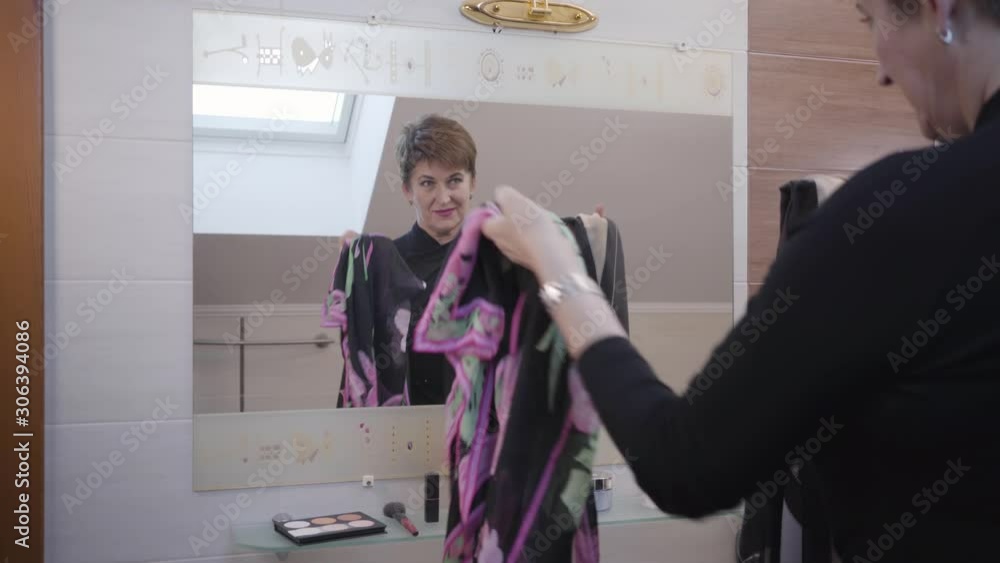 Mature Caucasian senior woman looking at two scarfs in front of big mirror in bathroom. Elegant businesswoman choosing outfit in the morning. Style, fashion, elegance. Mature Caucasian senior woman looking at two scarfs in front of big mirror in bathroom. Elegant businesswoman choosing outfit in the morning. Style, fashion, elegance.