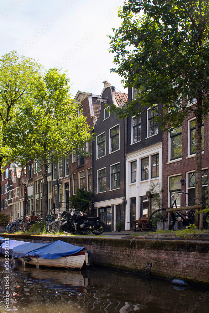 View of canal, trees, parked bicycles and boats, historical and traditional buildings showing Dutch architecture style in Amsterdam. It is sunny summer day.