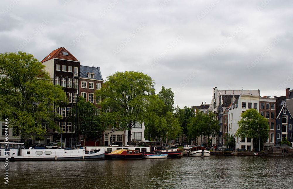 View of Amstel river, parked boats, trees, historical and traditional buildings in Amsterdam. It is a summer day with cloudy sky.