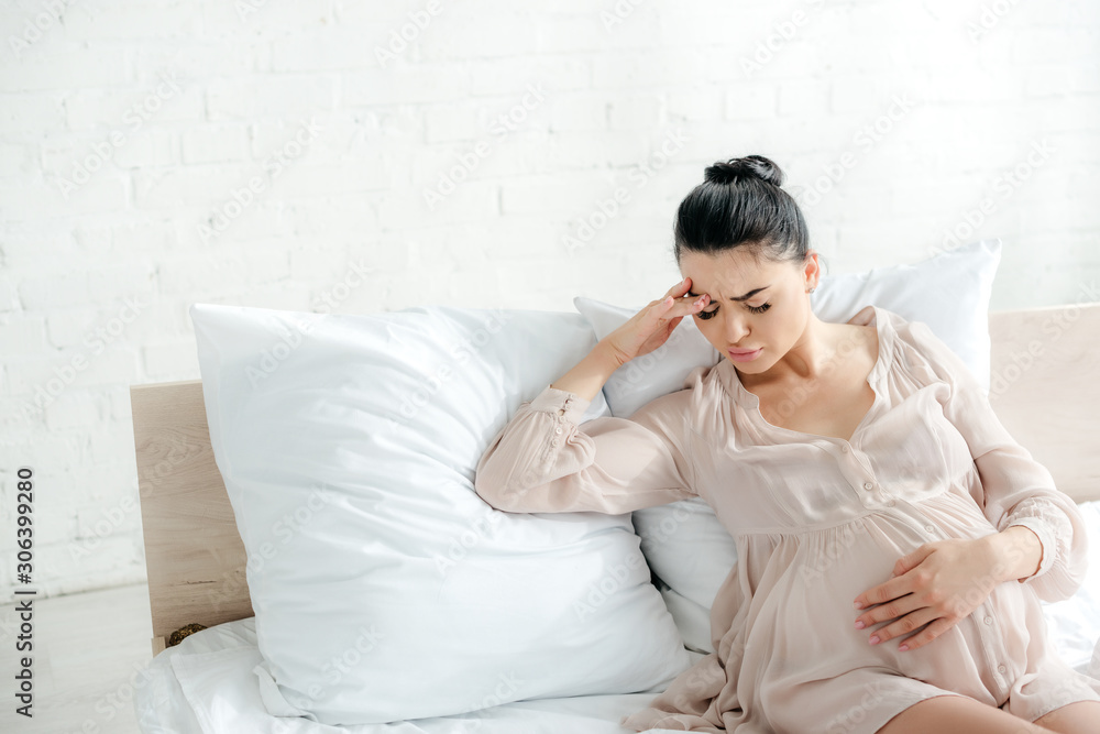 tired pregnant woman in nightie having headache and touching tummy while sitting on bed