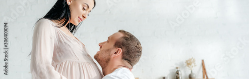 panoramic shot of handsome husband hugging his smiling pregnant wife in bedroom