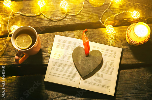 Holidays mood photo. Christmas lights and hot tea mug. Book for cosy evening. Sweet gingerbread and wooden heart on tray. Perfect winter flat lay. Hygge concept.