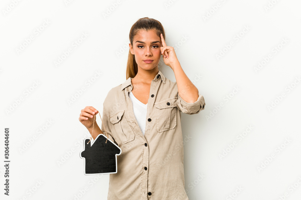 Young caucasian woman holding a house icon pointing his temple with finger, thinking, focused on a task. Young caucasian woman holding a house icon pointing his temple with finger, thinking, focused on a task.