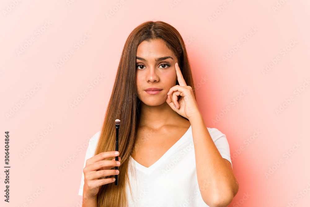 Fototapeta premium Young caucasian woman holding an skinbrush pointing his temple with finger, thinking, focused on a task.