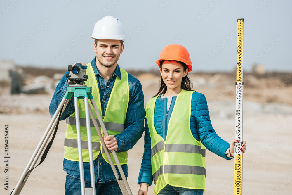 Smiling surveyors with ruler and digital level looking at camera - obrazy, fototapety, plakaty 