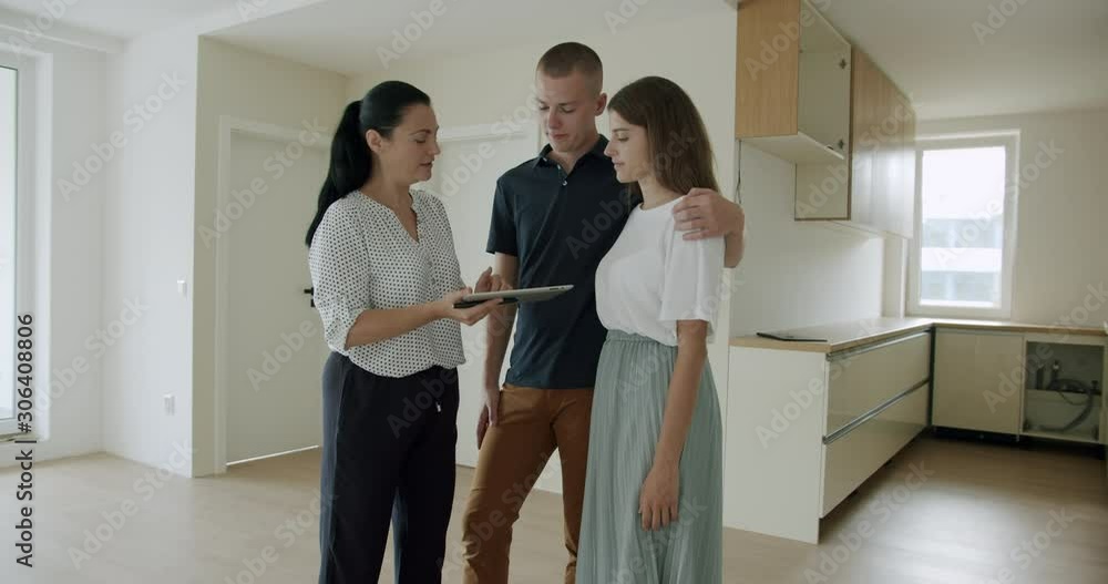 Estate agent showing young couple new apartment and using digital tablet. Female estate agent using digital tablet showing young couple in their 20s around new house. Estate agent showing young couple new apartment and using digital tablet. Female estate agent using digital tablet showing young couple in their 20s around new house.