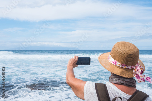Rear view of a senior woman at the sea in winter time taking a photo of the horizon over water. Casual clothing with backpack and hat. Cloudy sky