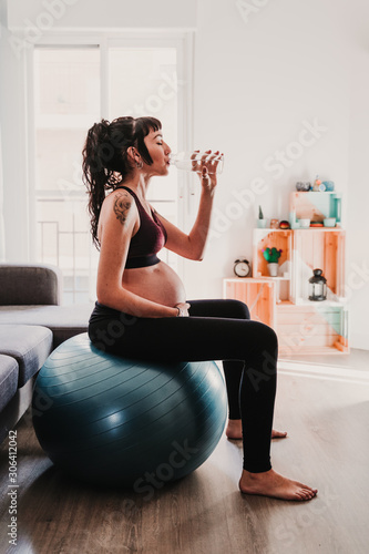 young pregnant woman at home sitting on pilates ball, drinking water. healthy lifestyle