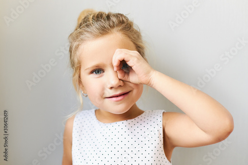 Horizontal shot of adorable pretty girl in beautiful dress posing in studio looking through binocular made of her hand, connecting thumb with fore finger. Cute funny female child having fun, spying