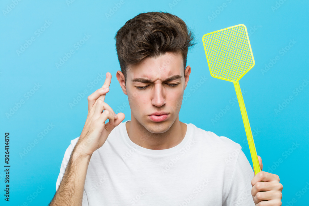 Young caucasian man holding a fly swatter crossing fingers for having luck Young caucasian man holding a fly swatter crossing fingers for having luck