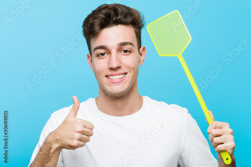 Fotografi Young caucasian man holding a fly swatter smiling and raising thumb up