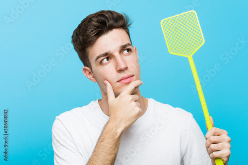 Fotografija Young caucasian man holding a fly swatter looking sideways with doubtful and skeptical expression