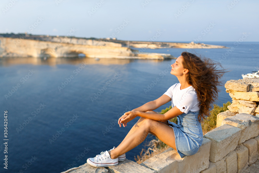 A happy woman sits on a rock at an altitude against the backdrop of the sea and inhales fresh sea air. Focus on the woman
