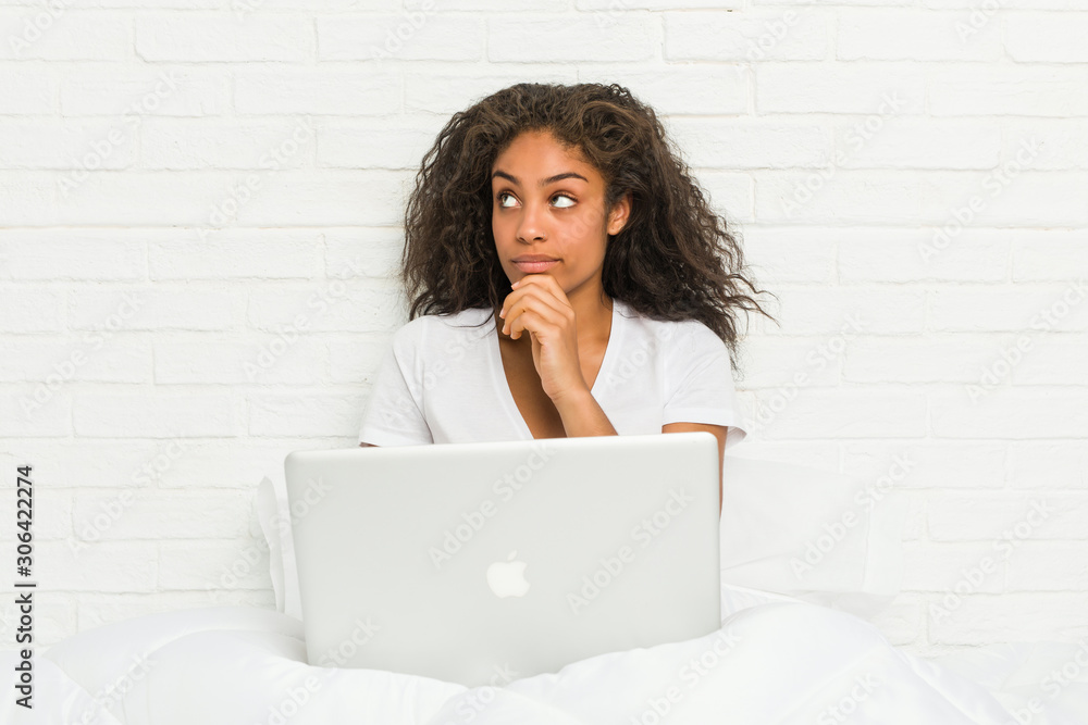 Young african american woman sitting on the bed with laptop looking sideways with doubtful and skeptical expression. Young african american woman sitting on the bed with laptop looking sideways with doubtful and skeptical expression.