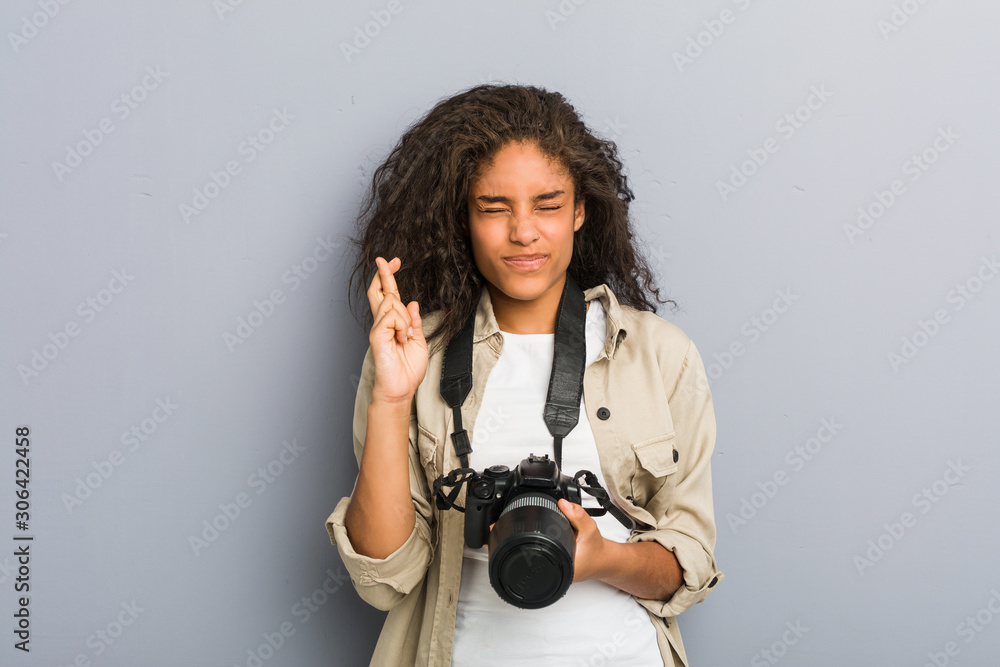 Fototapeta premium Young african american photographer woman holding a camera crossing fingers for having luck