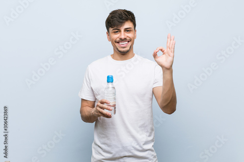 Young hispanic man holding a water bottle cheerful and confident showing ok gesture.