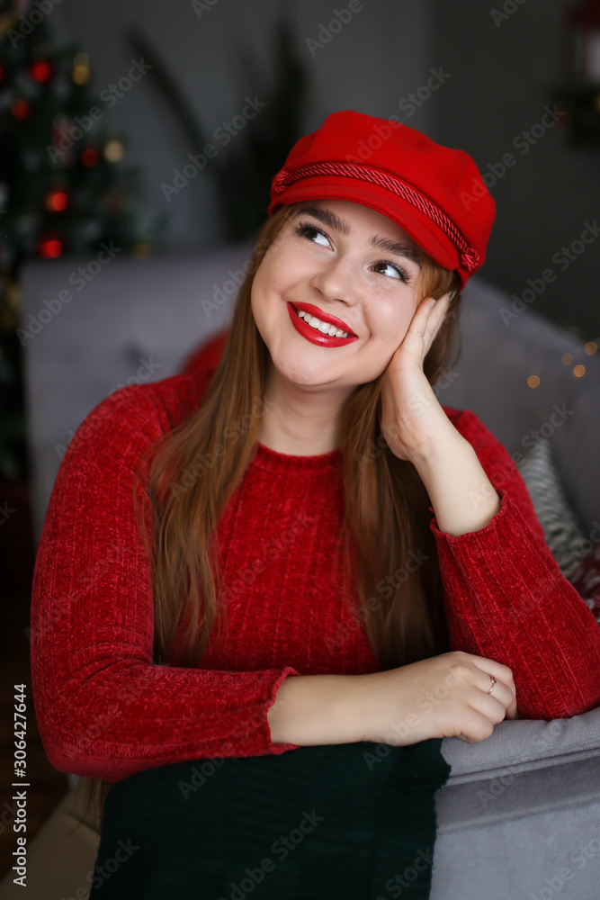 Beautiful bright attractive redhead girl plus size in a red sweater, beige leather skirt and red tights and a red cap rejoices and smiles on the background of the Christmas tree, gifts and bokeh. Beautiful bright attractive redhead girl plus size in a red sweater, beige leather skirt and red tights and a red cap rejoices and smiles on the background of the Christmas tree, gifts and bokeh.