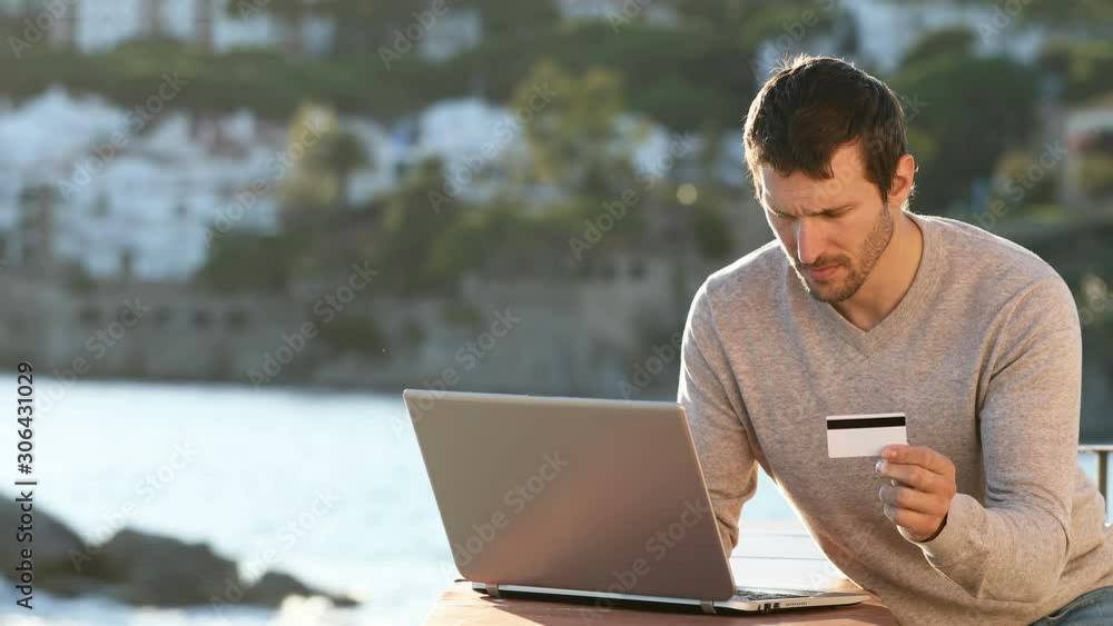 Serious man paying online in a balcony on the beach using a laptop and credit card Serious man paying online in a balcony on the beach using a laptop and credit card