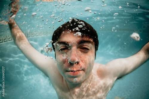 hombre joven caucásico de pelo moreno sumergido en una piscina, sacando burbujas de agua. apnea, nadar.