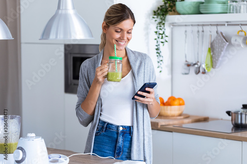 Pretty young woman using her mobile phone while drinking detox juice in the kitchen at home. photo