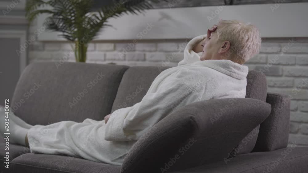Senior Caucasian woman lying on couch in white bathrobe. Mature lady having headache. Aging, healthcare, eternal love, care.