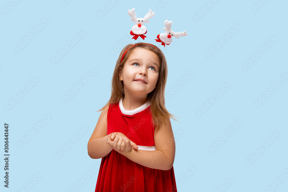 Cute Happy Child Smiles in a Red Dress, Girl wears a Christmas Santa Dress. Beautiful Kid is Laughing and posing in Studio. Child waving a Red dress. Little Girl at party. Isolated. Blue Background 
