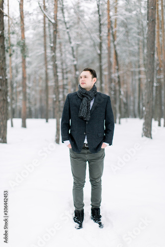 Handsome man in in stylish jacket and scarf walking in winter forest. Young groom outside in winter