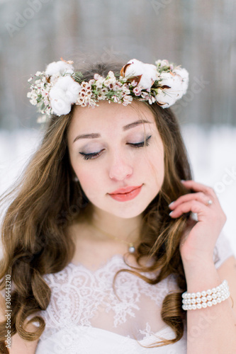 Beautiful bride touching her curly hair, with floral head band wreath, made from flowers and cotton, standing outdoors, on the background of winter forest