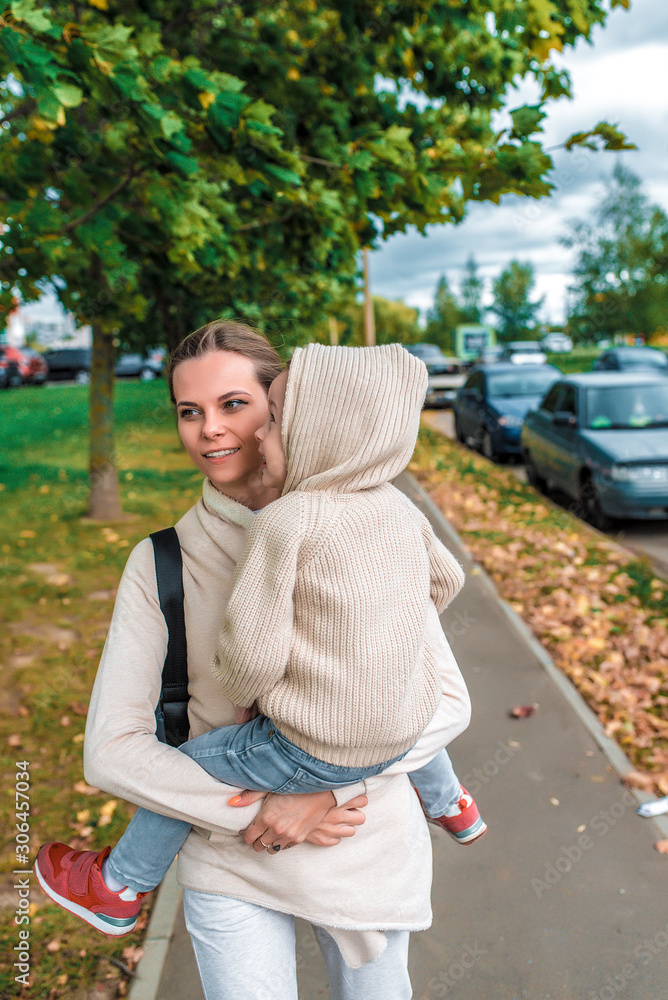 Woman mother walks down street, holds little boy son her arms, autumn city, walk along street, background trees, warm casual clothes, beige sweater with hood. Cold weather is breeze. Woman mother walks down street, holds little boy son her arms, autumn city, walk along street, background trees, warm casual clothes, beige sweater with hood. Cold weather is breeze.