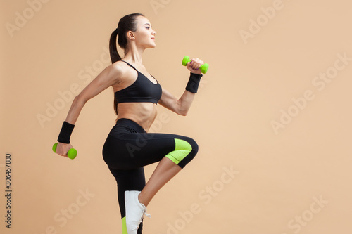 A young brunette woman is engaged in fitness, jumping with dumbbells, raising her knees high, on a peach background.