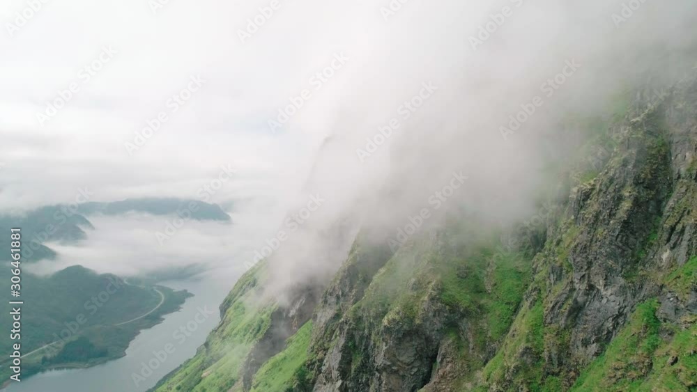 Stunning wide 4K drone shot with slow backward reveal motion through thick clouds climbing steep and majestic mountain range with dark rocky cliffs and ocean background view, Lofoten Nordland Norway. Stunning wide 4K drone shot with slow backward reveal motion through thick clouds climbing steep and majestic mountain range with dark rocky cliffs and ocean background view, Lofoten Nordland Norway.