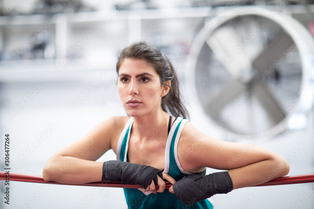 A badass female boxer is resting on ring ropes with her wraps on in a boxing gym, white walls, black and red mats, indoors. A badass female boxer is resting on ring ropes with her wraps on in a boxing gym, white walls, black and red mats, indoors.