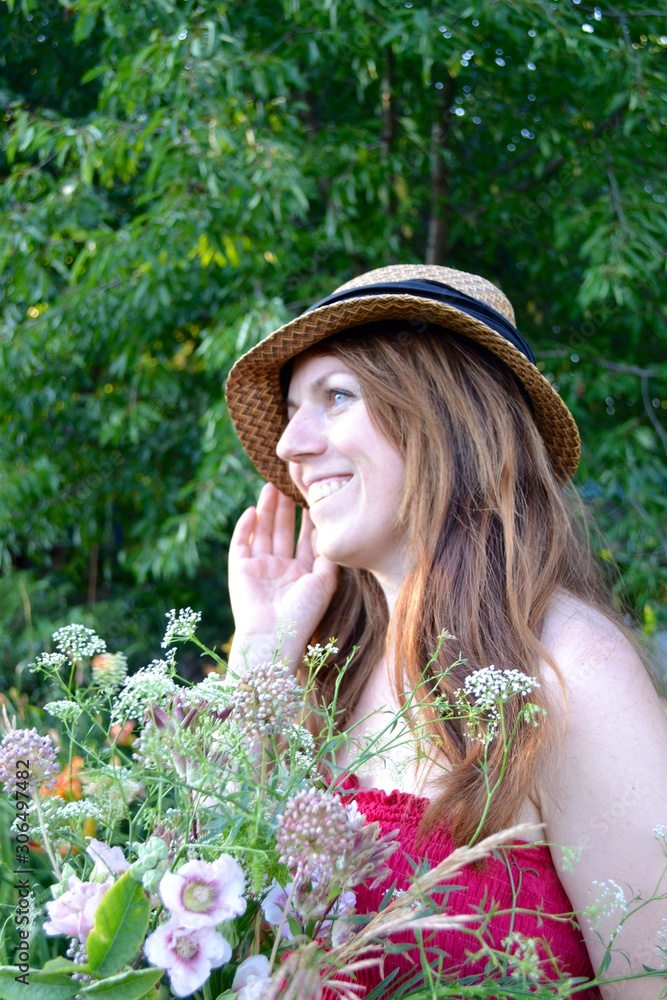 Fototapeta premium Portrait of a young woman with long brown hair in a hat and a raspberry sundress with a bouquet of wild flowers in a flowered summer garden