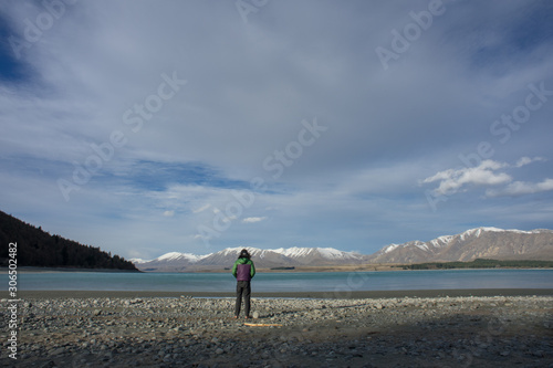 A man hiking by the lake shore with beatiful snow capped mountains in the background.View from behind.