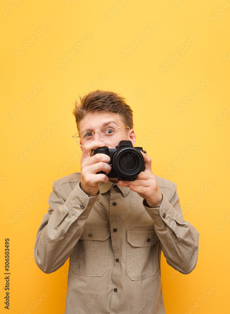 Portrait of funny photographer in shirt and glasses takes photo on camera against yellow background and looks into camera with funny face. Funny paparazzi takes a photo on camera. Isolated. Portrait of funny photographer in shirt and glasses takes photo on camera against yellow background and looks into camera with funny face. Funny paparazzi takes a photo on camera. Isolated.