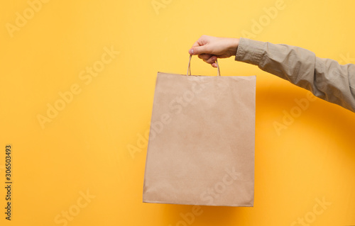 Human hands holds a paper bag in his hand on a yellow background. Kraft package holds man in shirt. Isolated. Ecological paper shopping bag in the hands of a man. Copy space