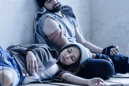 Homeless young man and woman are lying on cardboard on floor in abandoned house. Guy and girl addicts got overdose injections of heroin and getting high. Drug addiction and street life concept. photo
