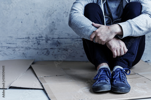 Unhappy guy is sitting on cardboard on floor in abandoned building. Homeless bum man lost everything because of abuse, addiction. Social problems in society. Loneliness and depression concept