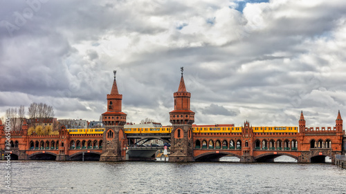 The famous bridge of bricks over the river Spree in Berlin is called Oberbaum Bridge. The picture is from south on a cloudy day. A yellow subway goes over the bridge.