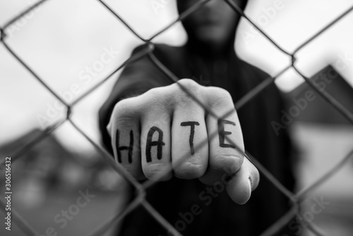 Aggressive teenage boy showing hes fist behind wired fence at the correctional institute, the word hate is written on hes hand, focus on the boys hand , conceptual image of juvenile delinquency .