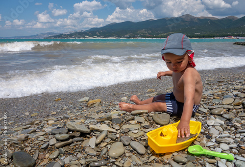 A little boy with panama hat plays on the beach by the sea with a yellow toy ship and green shoulder blade