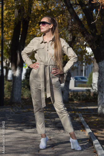 Attractive young woman in sunglasses and overalls similar to the uniform posing in autumn park with arms akimbo