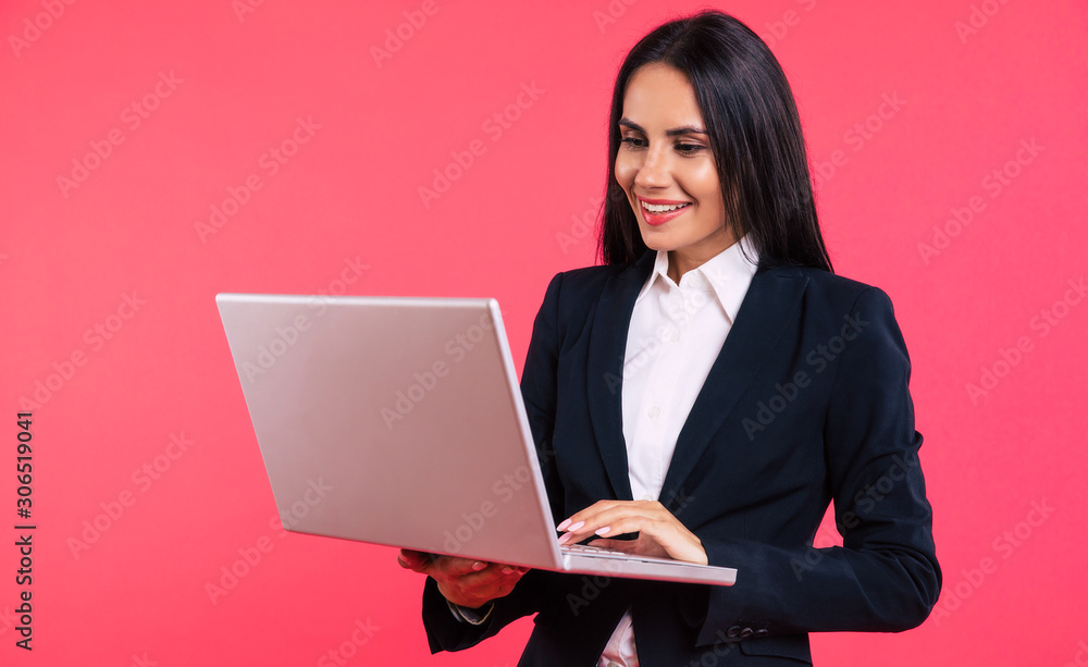 Creating projects. Close-up photo of a busy business woman in a smart outfit, who is smiling while holding a laptop in her arms and typing something or reading the information from the screen. Creating projects. Close-up photo of a busy business woman in a smart outfit, who is smiling while holding a laptop in her arms and typing something or reading the information from the screen.