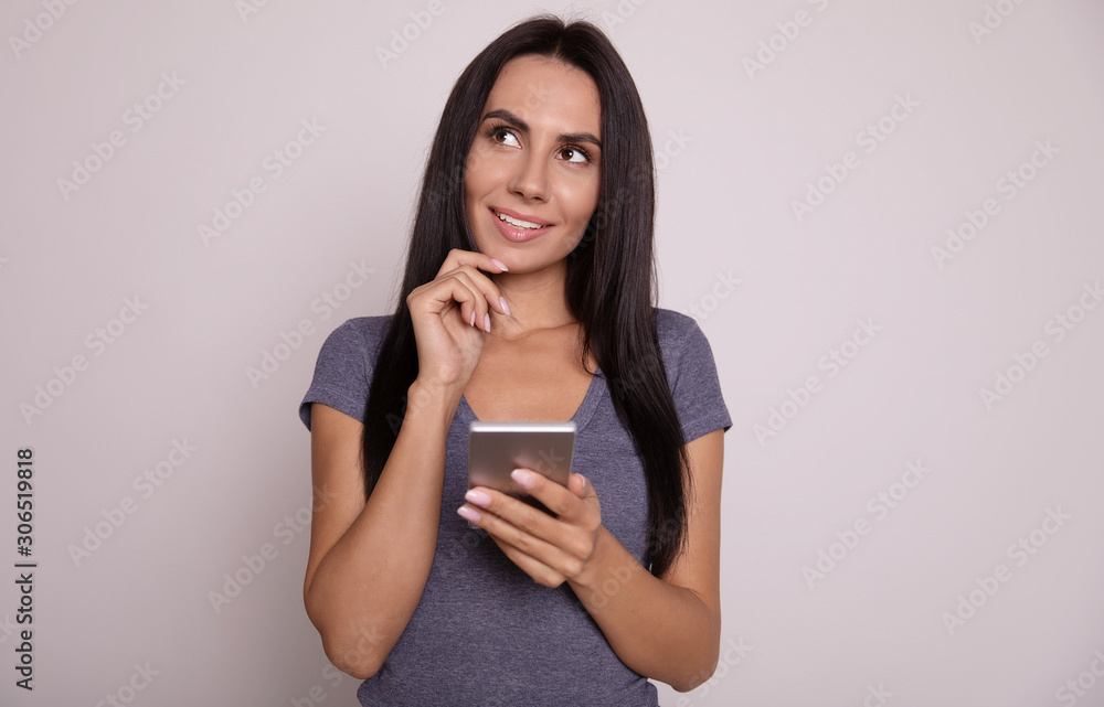 Thinking over the answer. Close-up photo of a stunning girl with straight black hair, who is looking to the left upper corner with a curious face expression, holding a smartphone in her right hand. Thinking over the answer. Close-up photo of a stunning girl with straight black hair, who is looking to the left upper corner with a curious face expression, holding a smartphone in her right hand.