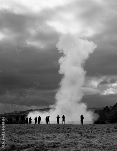 A massive geysir erupting among small shapes of tourists in wonder