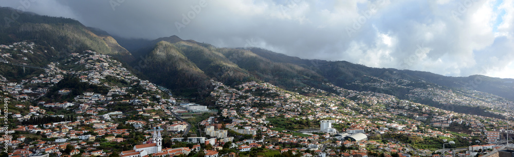 Wide panoramic view of Madeira island. Town with buildings on a steep mountain. Cloudy skies in background